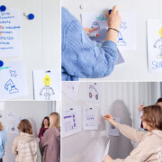 Eine Gruppe von Menschen steht an einem Whiteboard und befestigt und bespricht handgezeichnete Poster mit branchenbezogenen Symbolen und Notizen.