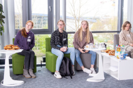 Vier junge Leute sitzen mit Snacks und Getränken auf Stühlen neben Tischen in einem hellen Raum mit großen Fenstern. Drei blicken nach links, einer nach vorne.