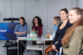 Fünf Frauen stehen und sitzen mit Wasserflaschen, Papieren und Snacks um einen Tisch in einer modernen Büroumgebung; im Hintergrund zeigt ein Bildschirm die Uhrzeit 09:00 Uhr an.