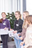Vier junge Menschen stehen im Haus und halten Zeichnungen von Fahrzeugen in den Händen. Der Blick ist nach vorne gerichtet. Im Hintergrund sind eine Pflanze und ein großes Fenster mit Jalousien zu sehen.