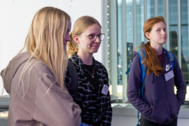 Drei junge Menschen stehen in einem Gebäude, in Freizeitkleidung und mit Namensschildern, mit Rucksäcken und einem Fenster mit vertikalen Jalousien im Hintergrund.