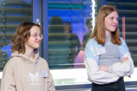Zwei junge Menschen, beide mit Namensschildern, stehen drinnen vor einem Fenster mit horizontalen Jalousien und blicken auf etwas außerhalb des Rahmens.