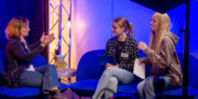 Drei Frauen sitzen auf blauen Stühlen und unterhalten sich in einem Studio. Zwei von ihnen halten Papiere und im Hintergrund sind Geräte zu sehen.