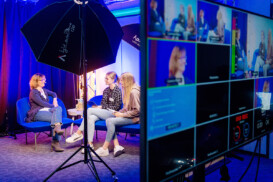 Drei Frauen sitzen und unterhalten sich an einem Set mit Studiobeleuchtung, während auf einem Monitor im Vordergrund Video-Feeds der aufgenommenen Szene angezeigt werden.