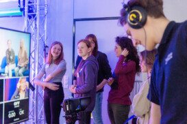 Eine Gruppe junger Leute steht in einem Studio, einige lächeln und unterhalten sich. Im Vordergrund sind ein großer Bildschirm und Kameraausrüstung zu sehen.