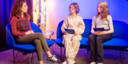 Drei Personen sitzen auf blauen Sofas in einem hell erleuchteten Raum, zwei Kinder halten Tablets in der Hand und stehen einem Erwachsenen während eines Gesprächs oder Interviews gegenüber.