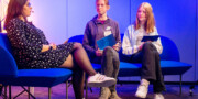 Drei Personen sitzen auf blauen Sofas in einem hell erleuchteten Raum; die Frau links spricht, während die beiden jungen Leute rechts zuhören und Ordner halten.