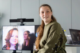 Eine Frau sitzt in einem Büro, im Hintergrund sind auf einem Monitor zwei Personen in einem Videoanruf zu sehen.