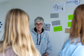 Eine Frau in einem grauen Kapuzenpullover steht vor einem Whiteboard mit verschiedenen Notizen und Diagrammen und spricht mit zwei Personen, deren Rücken zu sehen sind.
