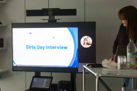 Eine Frau steht während eines Videoanrufs in einer Büroumgebung neben einem großen Bildschirm, auf dem der Text „Girls Day Interview“ angezeigt wird.