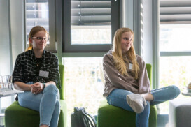 Zwei junge Frauen sitzen in Freizeitkleidung und mit Namensschildern auf grünen Stühlen in der Nähe eines Fensters und hören einer Veranstaltung im Innenbereich aufmerksam zu.