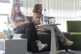 Zwei Personen sitzen mit Blick nach vorne auf einem grauen Sofa, vor ihnen stehen Getränkeflaschen und Gläser auf einem kleinen weißen Tisch in einem hellen Raum mit vertikalen Jalousien.