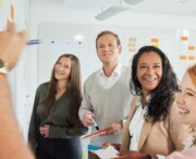 Five people stand by a whiteboard with sticky notes, collaborating and smiling, as one person writes or points at the board.
