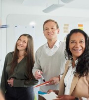 Five people stand by a whiteboard with sticky notes, collaborating and smiling, as one person writes or points at the board.