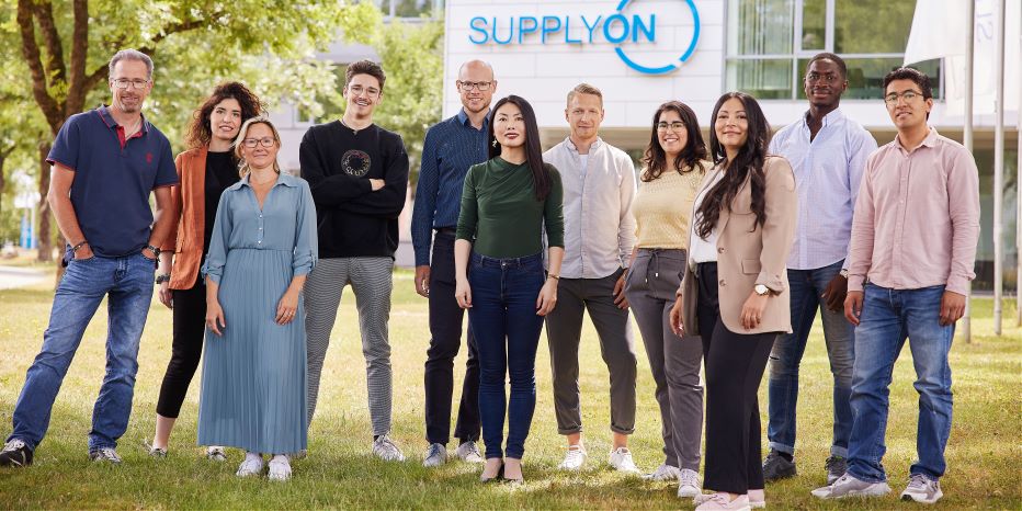 A group of eleven people stands outdoors in front of a building with the sign SUPPLYON, posing for a group photo on a sunny day.