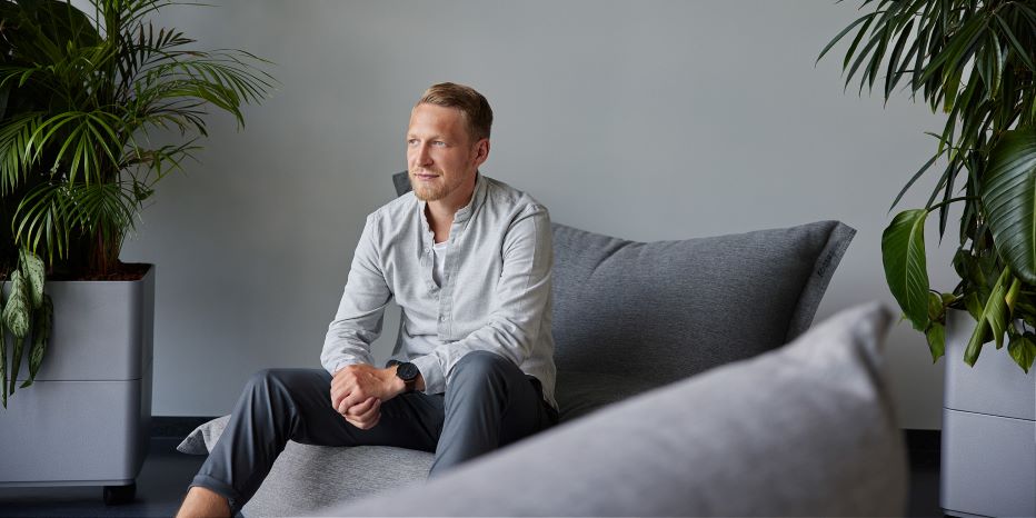 A man sits on a gray sofa in a modern room with potted plants, wearing a light shirt and dark pants, looking slightly to the side.