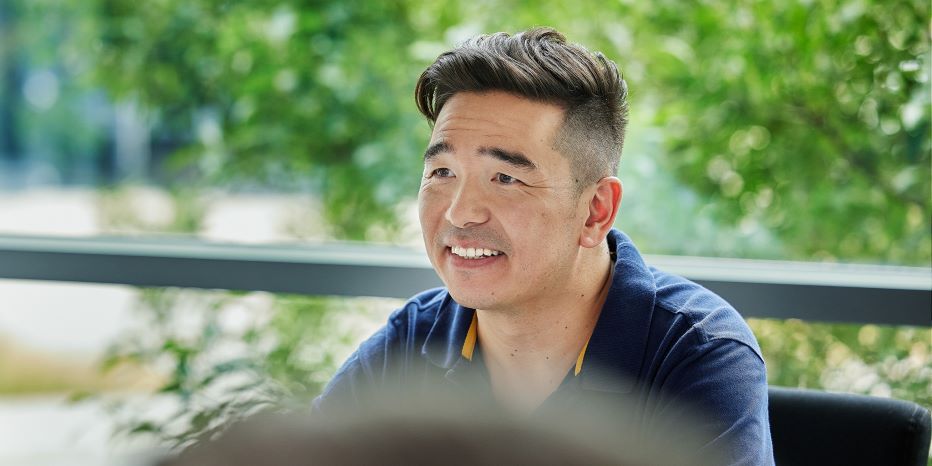 A man with short dark hair, wearing a navy shirt, sits indoors near a window with greenery outside, smiling and looking slightly to the side.
