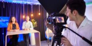 A person operates a camera recording two people standing behind a table in a brightly lit studio with a screen and decorative lights.