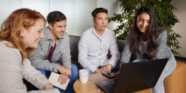Four people sit together on a sofa, looking at a laptop and smiling, with a plant and mugs on the table in front of them.