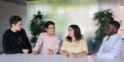 Four people sit at a white table having a conversation in a modern office with large windows and green plants, discussing opportunities for a Werkstudent Supply Chain position.