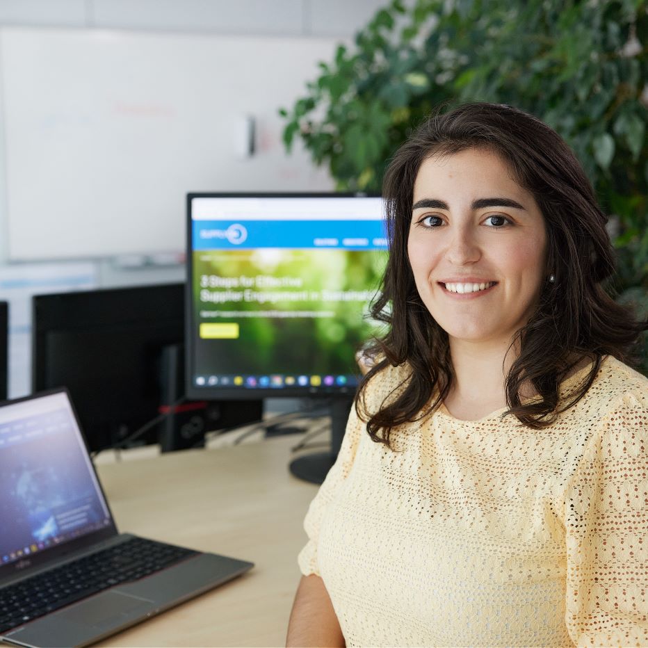 A young professional woman sits at a desk with a laptop open in front of her. A computer monitor behind her displays a website, while a large plant adds greenery to the workspace.