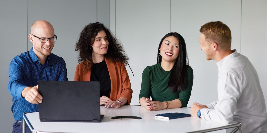 Four people sit around a table, engaged in conversation, with one person using a laptop.