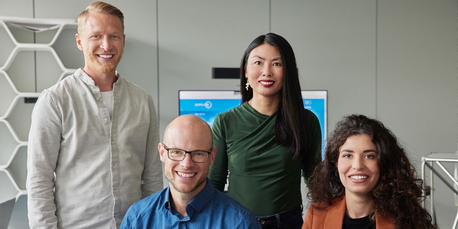 Four people pose indoors in front of a monitor, with two standing and two sitting, dressed in business-casual attire.