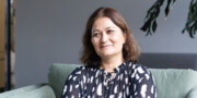 A woman with shoulder-length brown hair sits on a green sofa, wearing a black and white patterned blouse, her slight smile reflecting the confidence of someone embracing Agile Learning.
