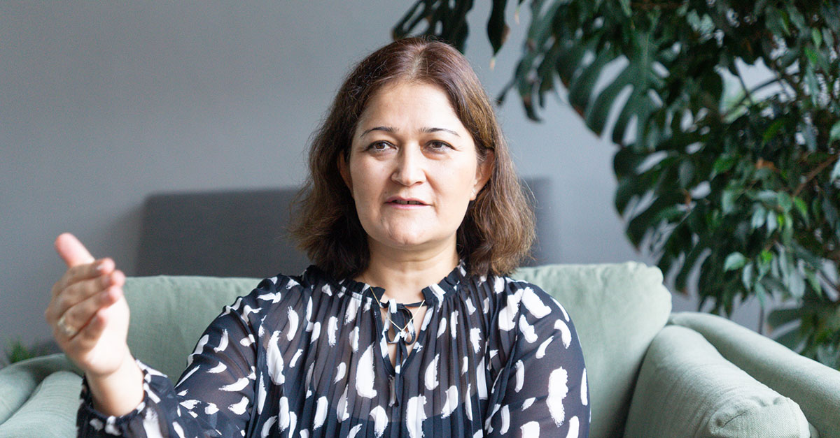 A woman with shoulder-length brown hair sits on a green sofa, gesturing with her right hand as she discusses Agile Learning, surrounded by leafy plants in the background.