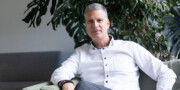 A man with short gray hair wearing a white shirt sits on a green chair indoors, a large leafy plant in the background symbolizing Gesundheit und Erfolg.