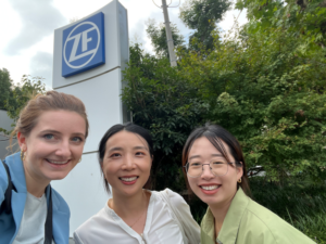 Three women stand outdoors in front of a ZF company sign in Shanghai, smiling for a photo with greenery and trees in the background.