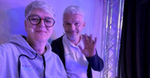 Two people, one in glasses and a hoodie, the other in a white shirt, stand indoors under purple lighting. The person on the right is waving, perhaps greeting a supplier at an event.