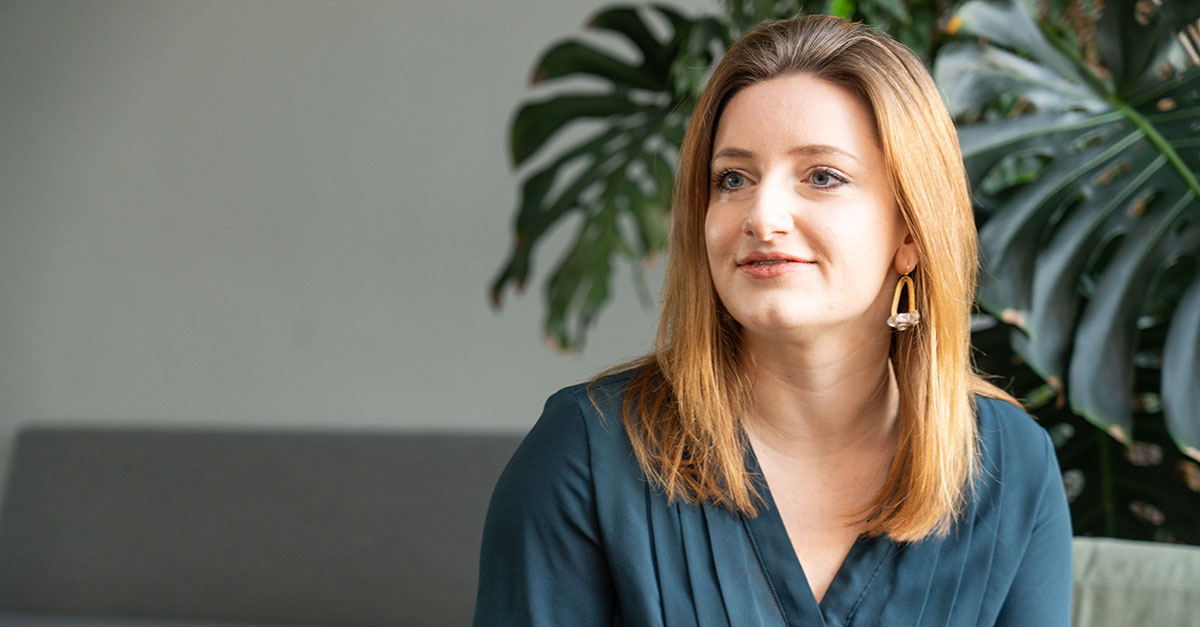 A Supply Chain Digitalisierung Consultant sits indoors, her shoulder-length light brown hair complementing a dark green blouse and gold earrings, framed by a large leafy plant as she reflects on ihre Aufgaben.