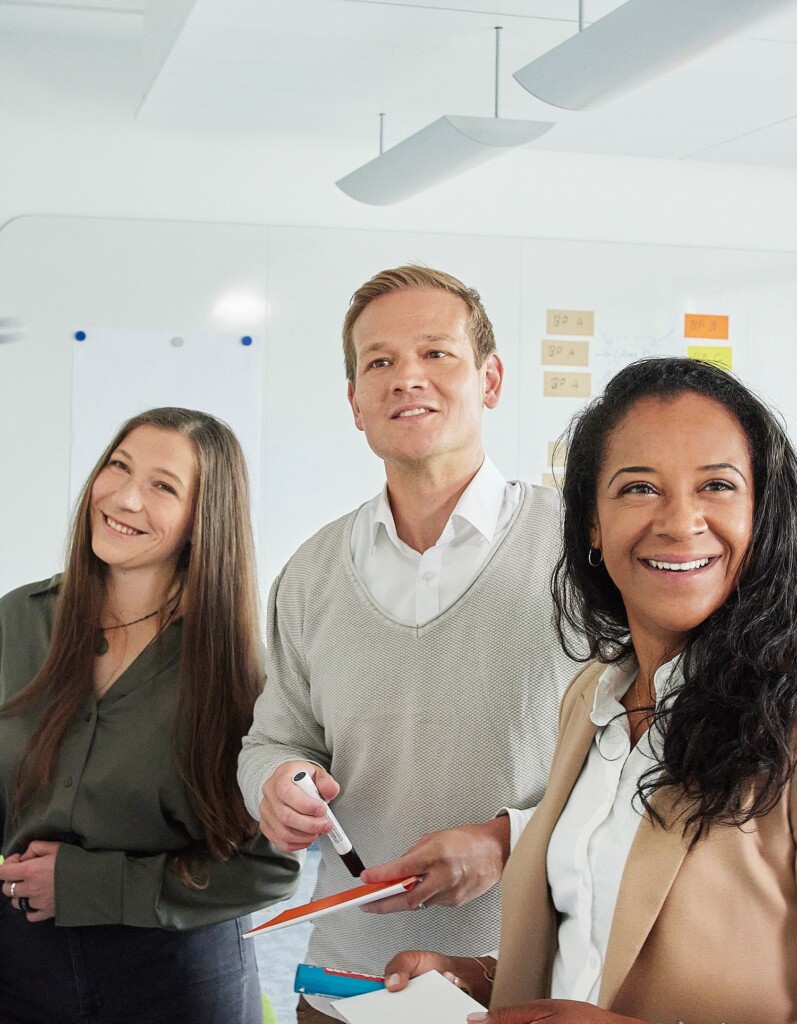 Three professionals stand in an office setting, holding markers and notepads, smiling and looking towards something off-camera.