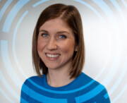 Woman with straight brown hair wearing a blue top, smiling at the camera; background features circular blue graphic patterns on a white gradient.