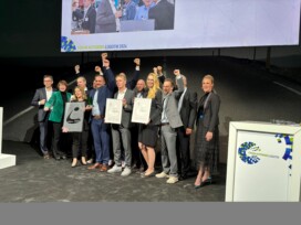 A group of people stand together on a stage, some raising their fists, holding certificates and an award at the Forum Automobillogistik 2024 event.