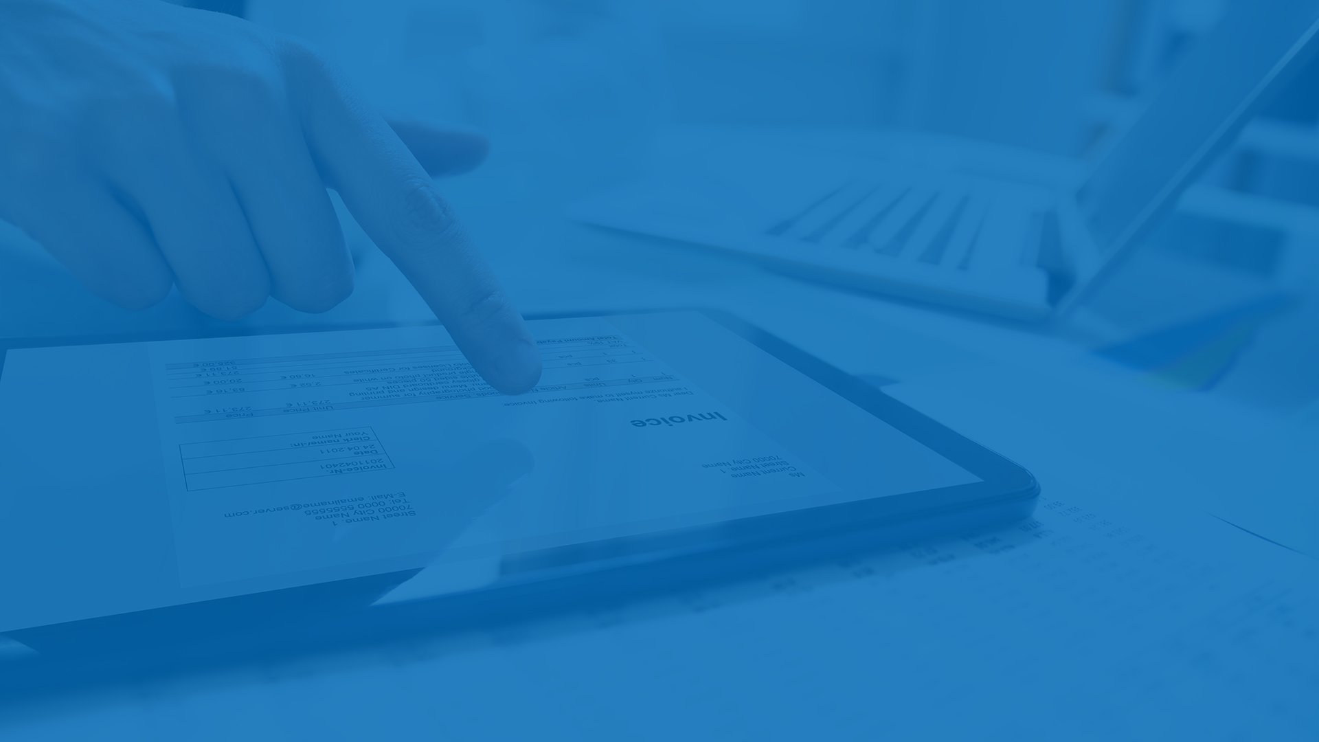 A person points to an invoice displayed on a tablet, with a laptop and documents visible in the background.