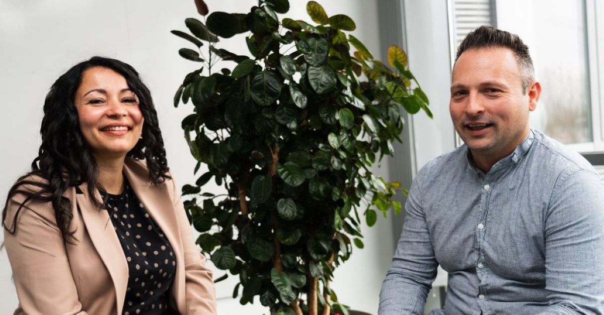 Two people sit on either side of a leafy indoor plant, smiling at the camera in a bright, modern room—an inviting space perfect for interactive team meeting design.