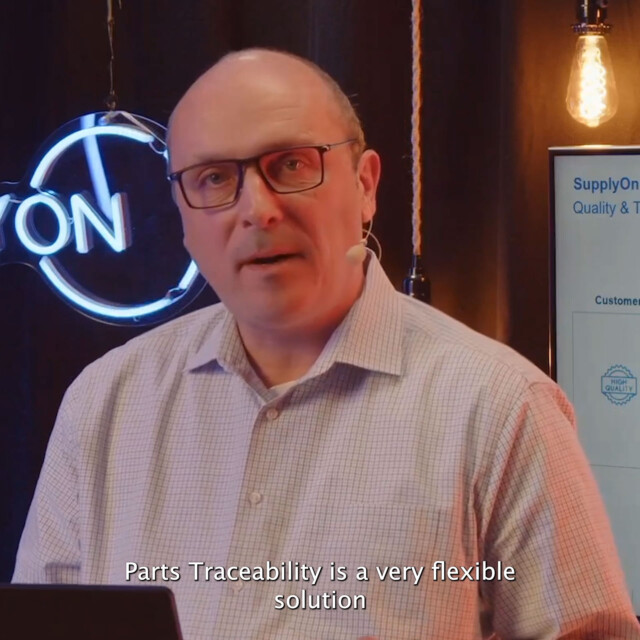 A man wearing glasses and a checked shirt speaks in front of a laptop about parts traceability; a neon sign and a presentation screen are in the background. Subtitles read: Parts Traceability is a very flexible solution.
