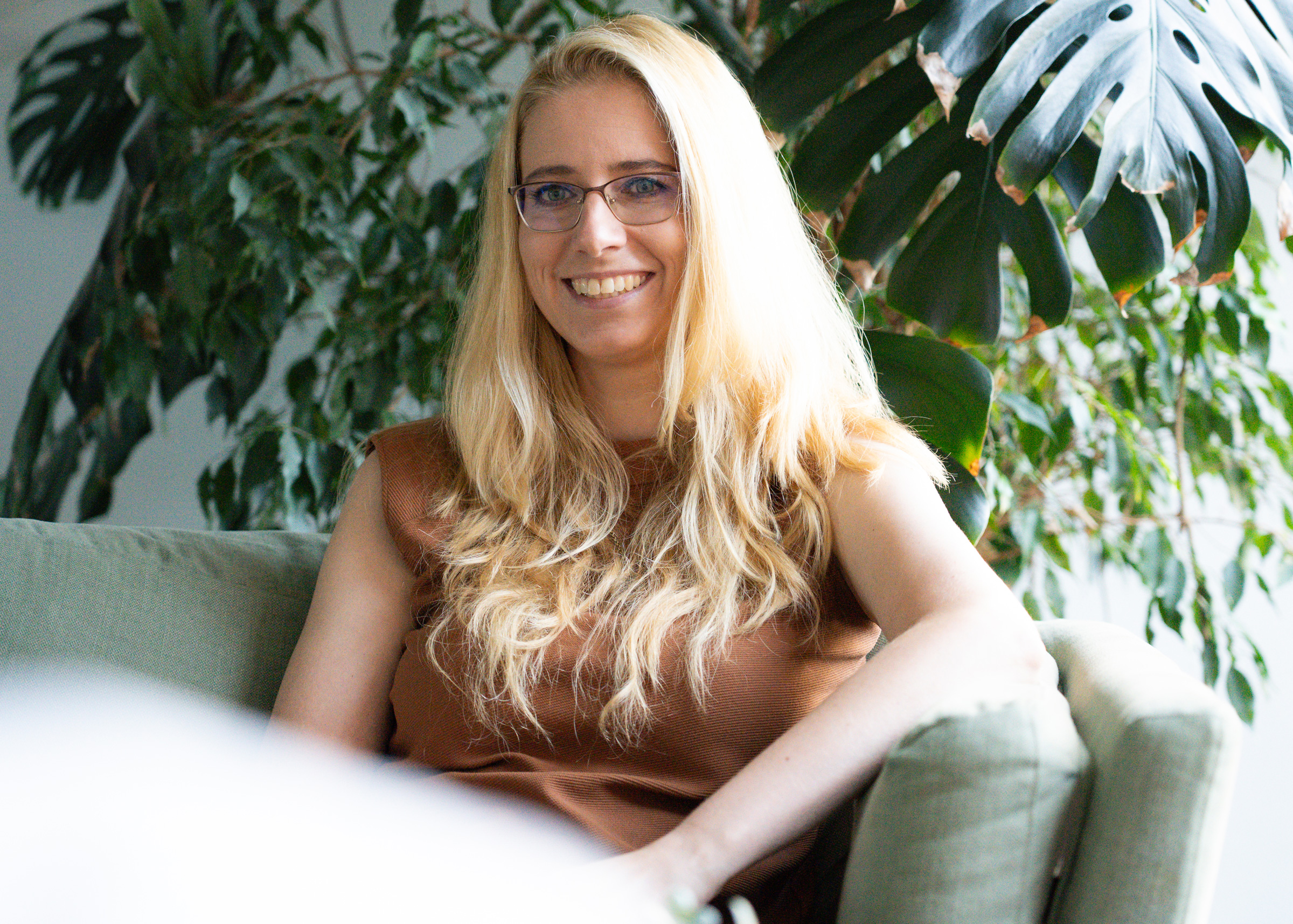A woman with long blonde hair and glasses sits on a light green couch, smiling, with leafy green plants in the background—radiating a sense of positivity and embracing a positive error culture.