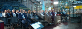 A group of people in business attire sit in rows of chairs at an indoor conference or presentation, with screens and industrial equipment visible in the background.