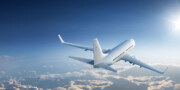 A commercial airplane flies above the clouds under a bright sun in a clear blue sky.