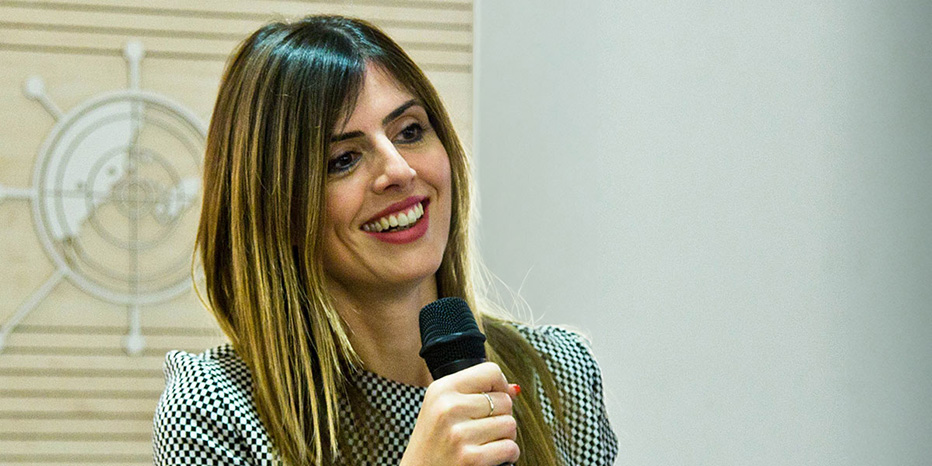 A woman with long hair holds a microphone and smiles while speaking about MRO indoors, with a patterned background and plain wall visible.