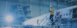 An MRO engineer wearing a hard hat and safety vest walks down stairs beside an airplane inside an aircraft maintenance hangar.