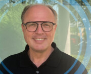 A middle-aged man with glasses and a black collared shirt smiles at the camera, standing in front of a window with blue circular graphics overlaid.