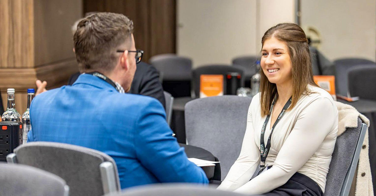 Two people sit across from each other at a table in a conference room, engaged in conversation about the pharmaceutical supply chain. Both wear event lanyards, and the woman is smiling.