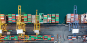 Aerial view of shipping containers and cranes at a port terminal, with cargo stacked and machinery positioned along the dock.
