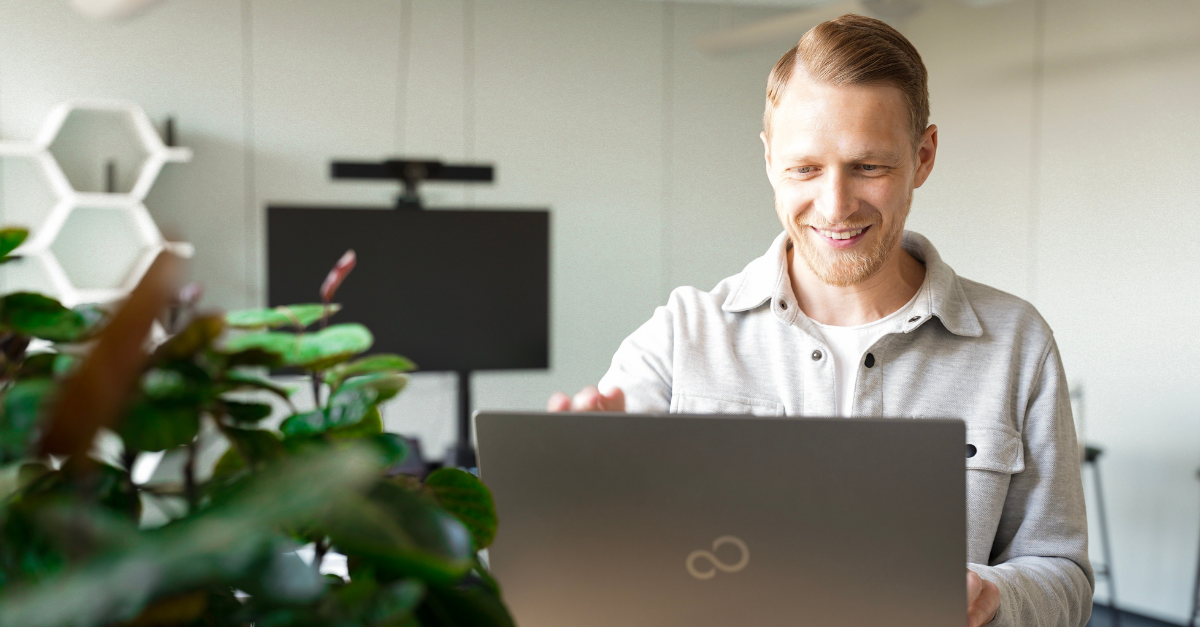 A person smiling while using a laptop in a bright office, embodying leadership in digitalisation, with a plant in the foreground and a monitor in the background.