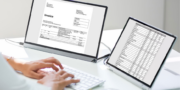 A person types on a keyboard at a desk with two monitors displaying an invoice and a spreadsheet.