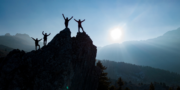Four people stand on rocky peaks with arms raised in silhouette, backlit by the sun, with mountains and trees visible in the background.